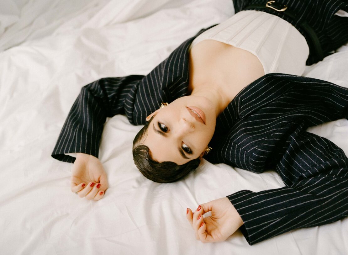A woman with dark hair in a striped blazer lies on a white sheet, looking at the camera with a neutral expression indoors.