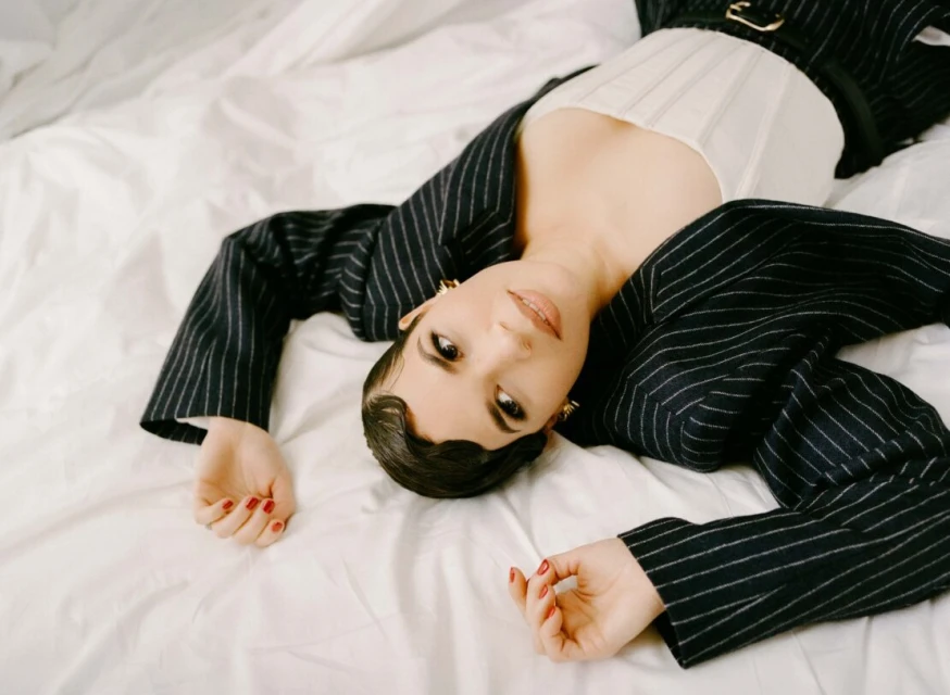 A woman with dark hair in a striped blazer lies on a white sheet, looking at the camera with a neutral expression indoors.