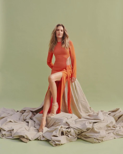 A woman with long brown hair poses in an orange dress with strappy heels, sitting on a tan textile against a light green backdrop.