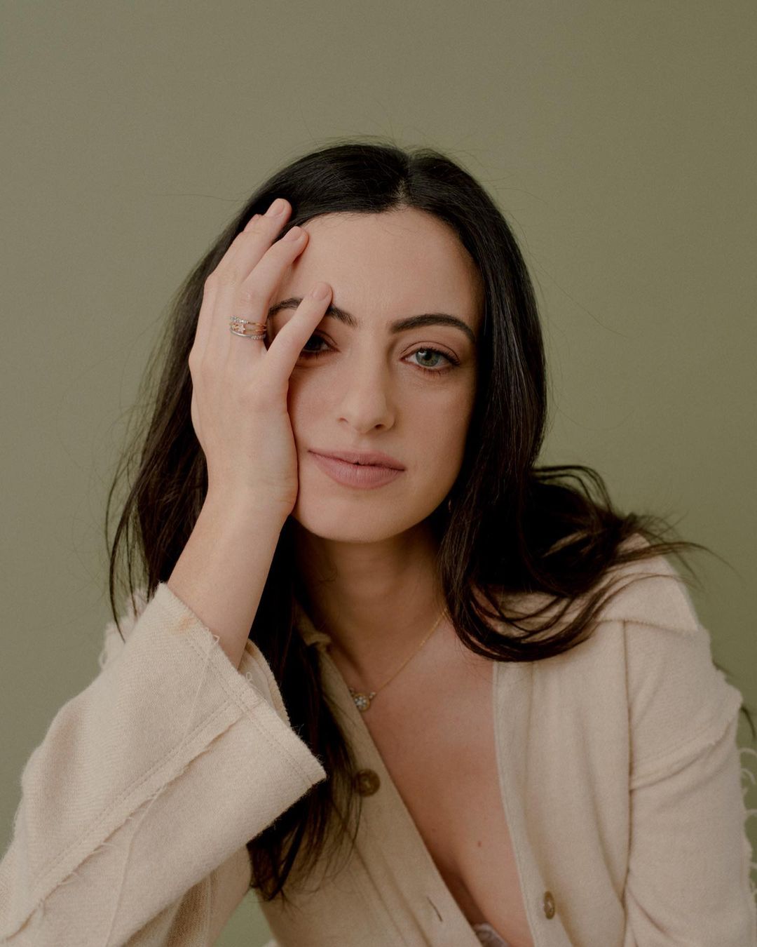 Portrait of a young woman with dark hair, wearing a beige jacket, resting her face on her hand against a muted green backdrop.