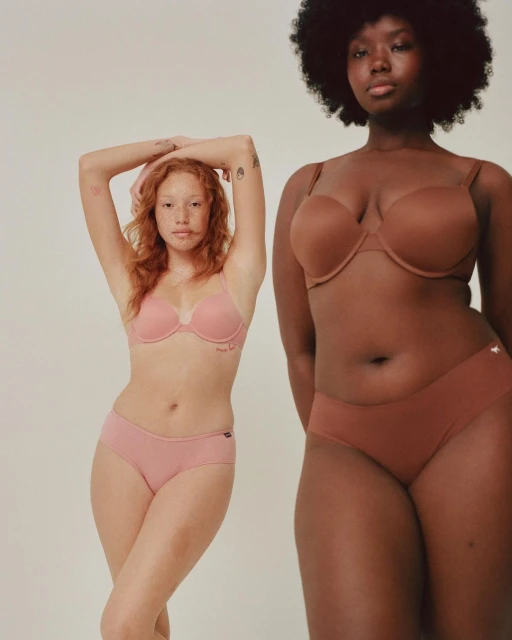 Two women pose in a photo studio wearing matching bra and panty sets, showcasing light pink and brown tones against a neutral background.