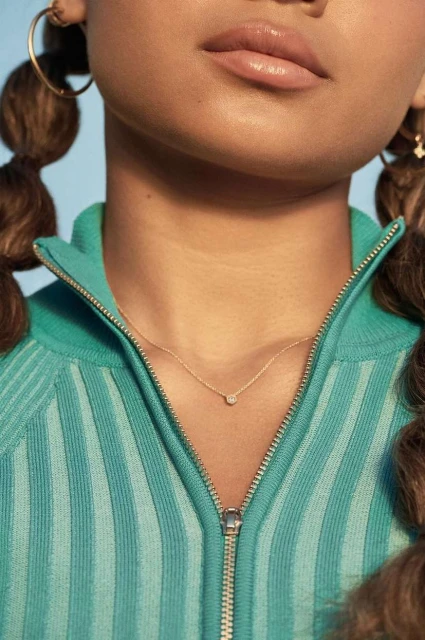 Young woman with brown hair in pigtails wearing a turquoise ribbed zip-up top and delicate gold necklace with small diamond pendant.