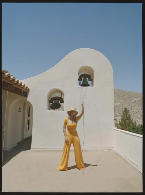A woman in a yellow jumpsuit and hat rings church bells in a Spanish-style setting under a clear blue sky. Outdoor scene.