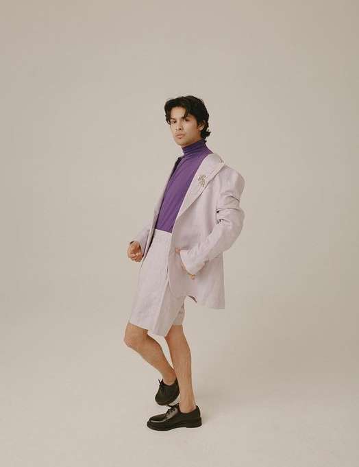 Fashion portrait of a young male adult in a lilac suit and purple turtleneck, posing in a photo studio against a light beige background.
