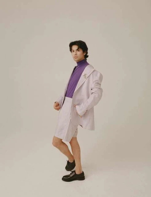 Fashion portrait of a young male adult in a lilac suit and purple turtleneck, posing in a photo studio against a light beige background.