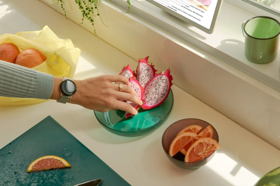 Close-up of a woman's hand selecting dragon fruit from a glass bowl on a kitchen counter with oranges and a digital tablet nearby.