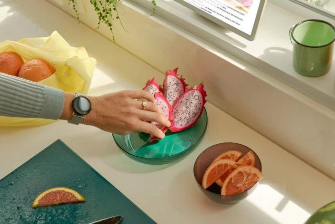 Close-up of a woman's hand selecting dragon fruit from a glass bowl on a kitchen counter with oranges and a digital tablet nearby.