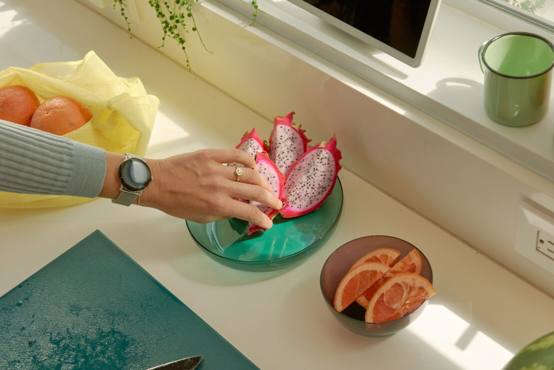 Overhead shot of a person's hand reaching for a dragon fruit in a bowl next to grapefruit slices on a white countertop indoors.