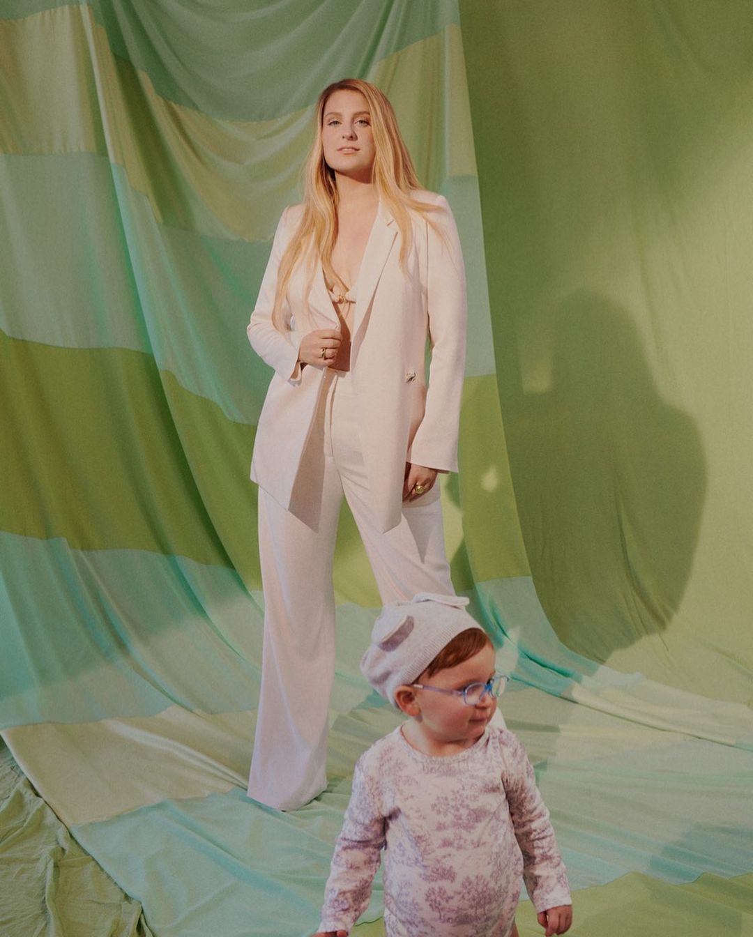 A woman in a pantsuit stands next to a baby wearing a bonnet and glasses in a studio with a green draped backdrop, full shot.