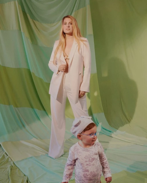 A woman in a pantsuit stands next to a baby wearing a bonnet and glasses in a studio with a green draped backdrop, full shot.