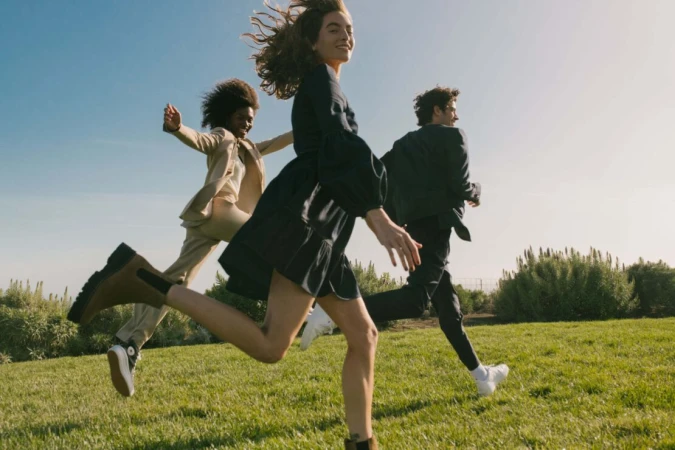Three models running across a green grassy field under a clear blue sky, captured in a dynamic low-angle outdoor shot.