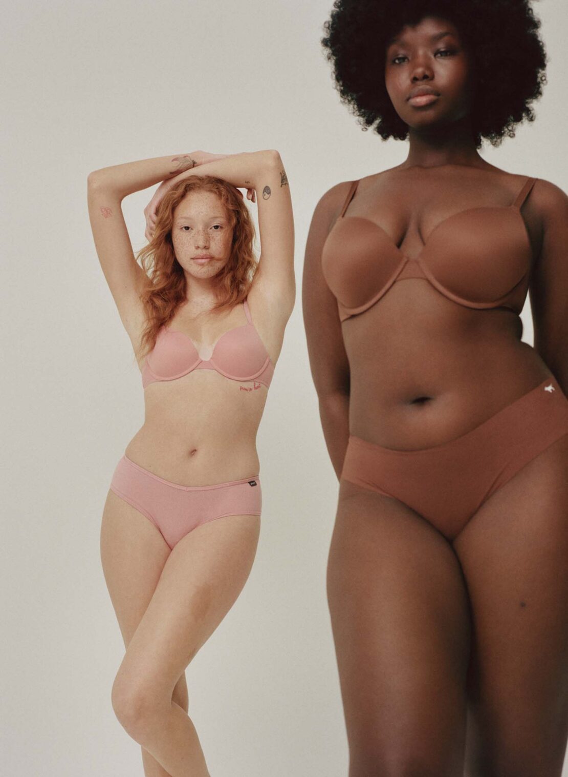 Two women of different skin tones pose in matching bras and underwear in a studio shot against a neutral background.