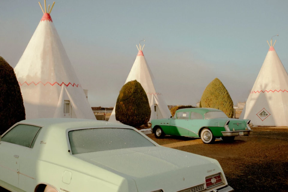 Cars parked near teepee-shaped motel rooms on a sunny day. Vintage turquoise car and white sedan in an outdoor setting.