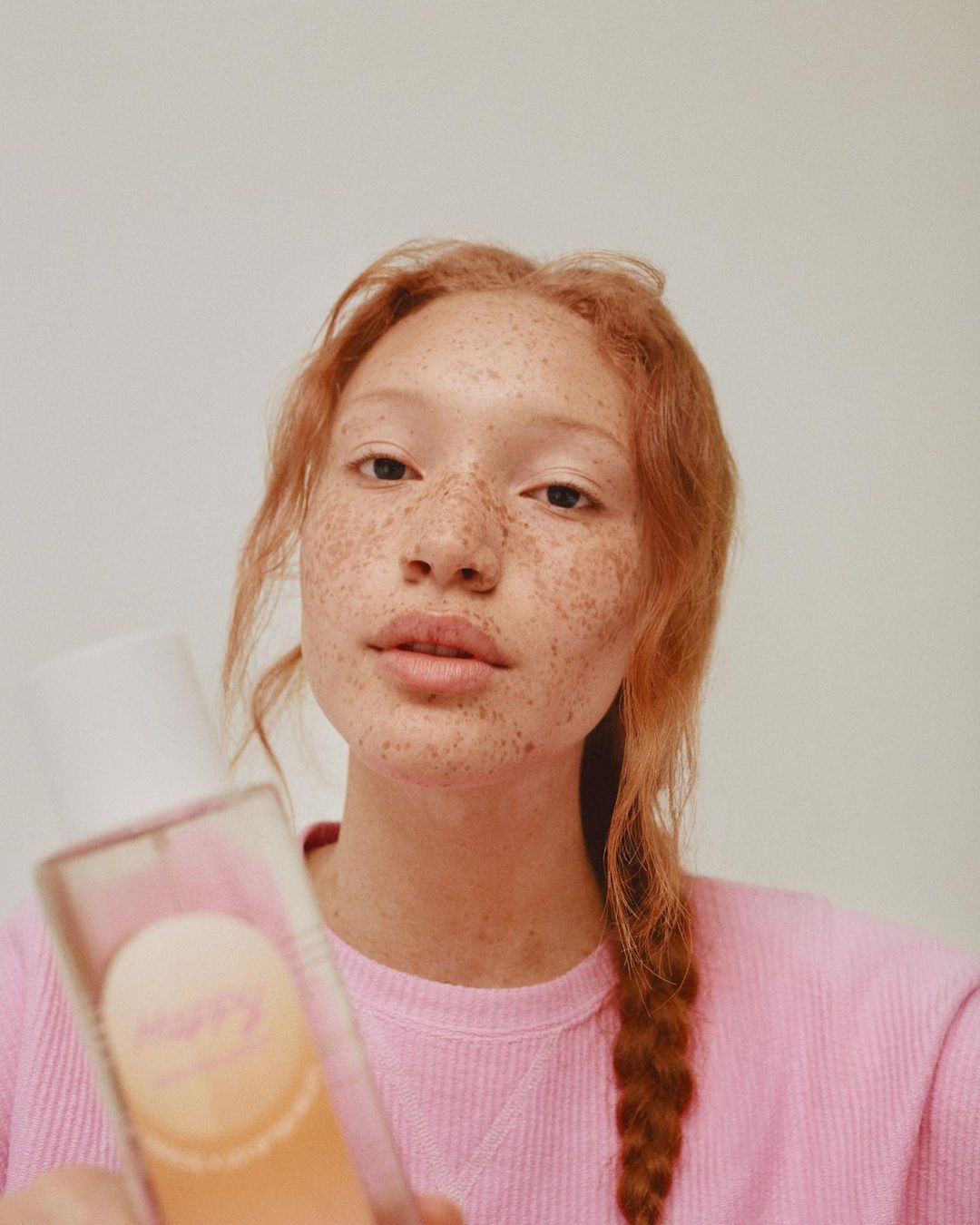 Close-up of a red-haired woman with freckles in a pink top, holding a bottle against a neutral background. Beauty and skincare focus.