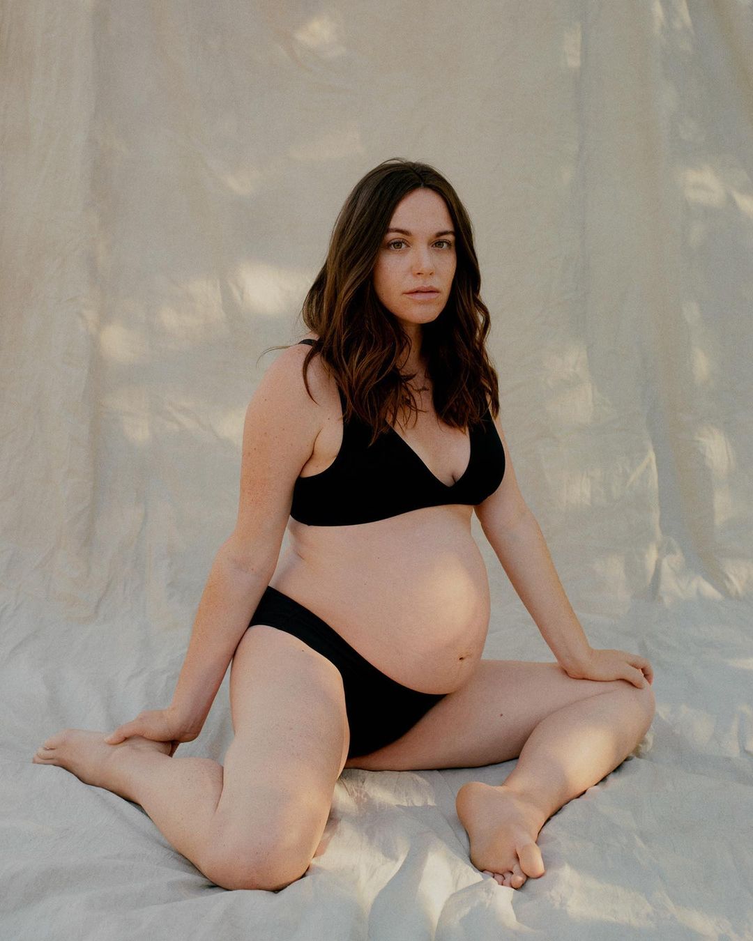 Portrait of pregnant woman in black bra and underwear sitting on cream fabric, showcasing maternity and body positivity in soft light.