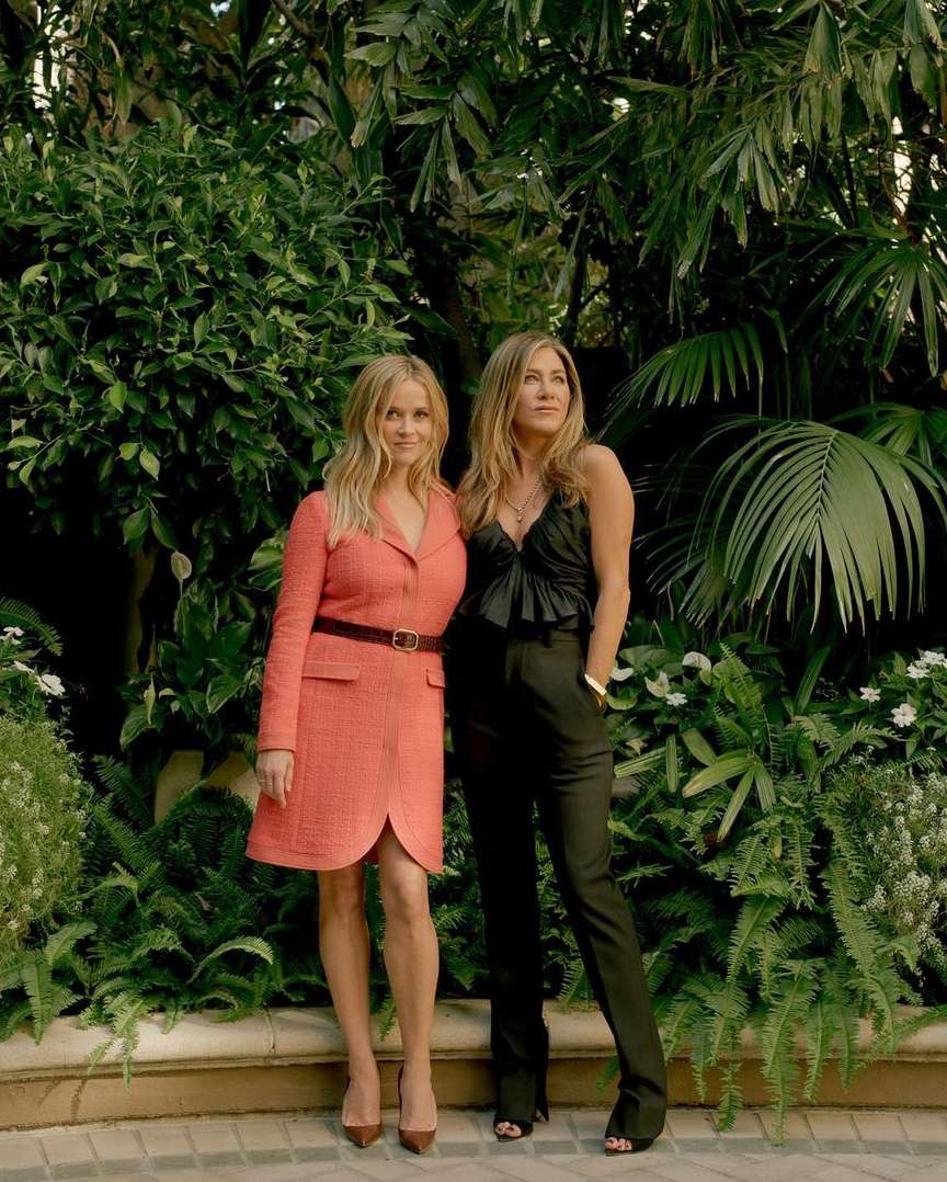 Two women, Jennifer Aniston and Reese Witherspoon, pose outdoors against a lush green backdrop, showcasing fashion and style.