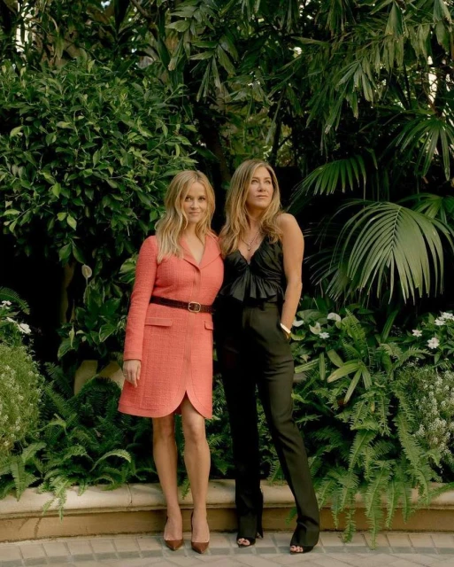 Two women, Jennifer Aniston and Reese Witherspoon, pose outdoors against a lush green backdrop, showcasing fashion and style.