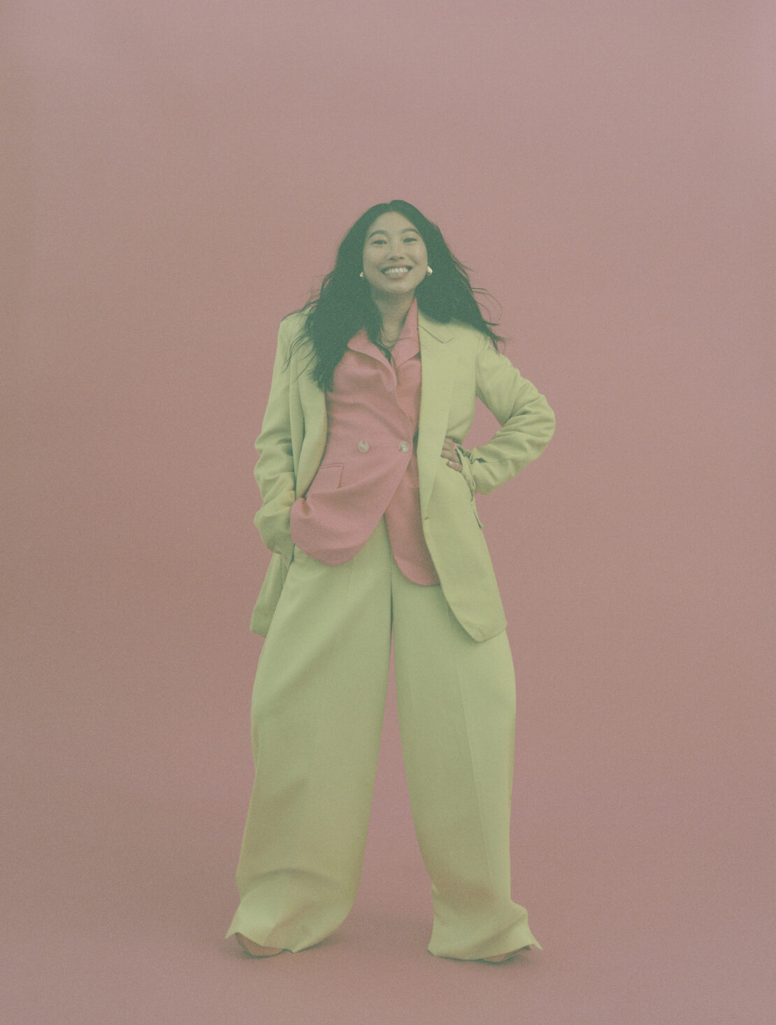 Smiling woman in green suit and pink shirt posing against light pink background in studio with hands in pockets, looking at camera.