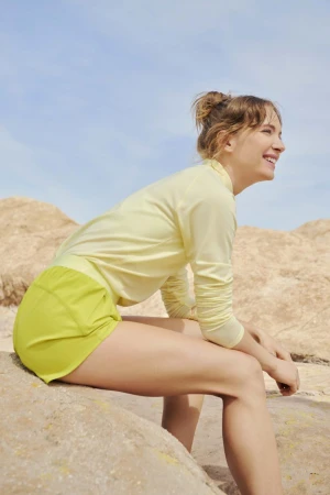 Woman in pale yellow long-sleeve top and shorts sitting outdoors on rocks under a clear blue sky, photographed by Amy Harrity.