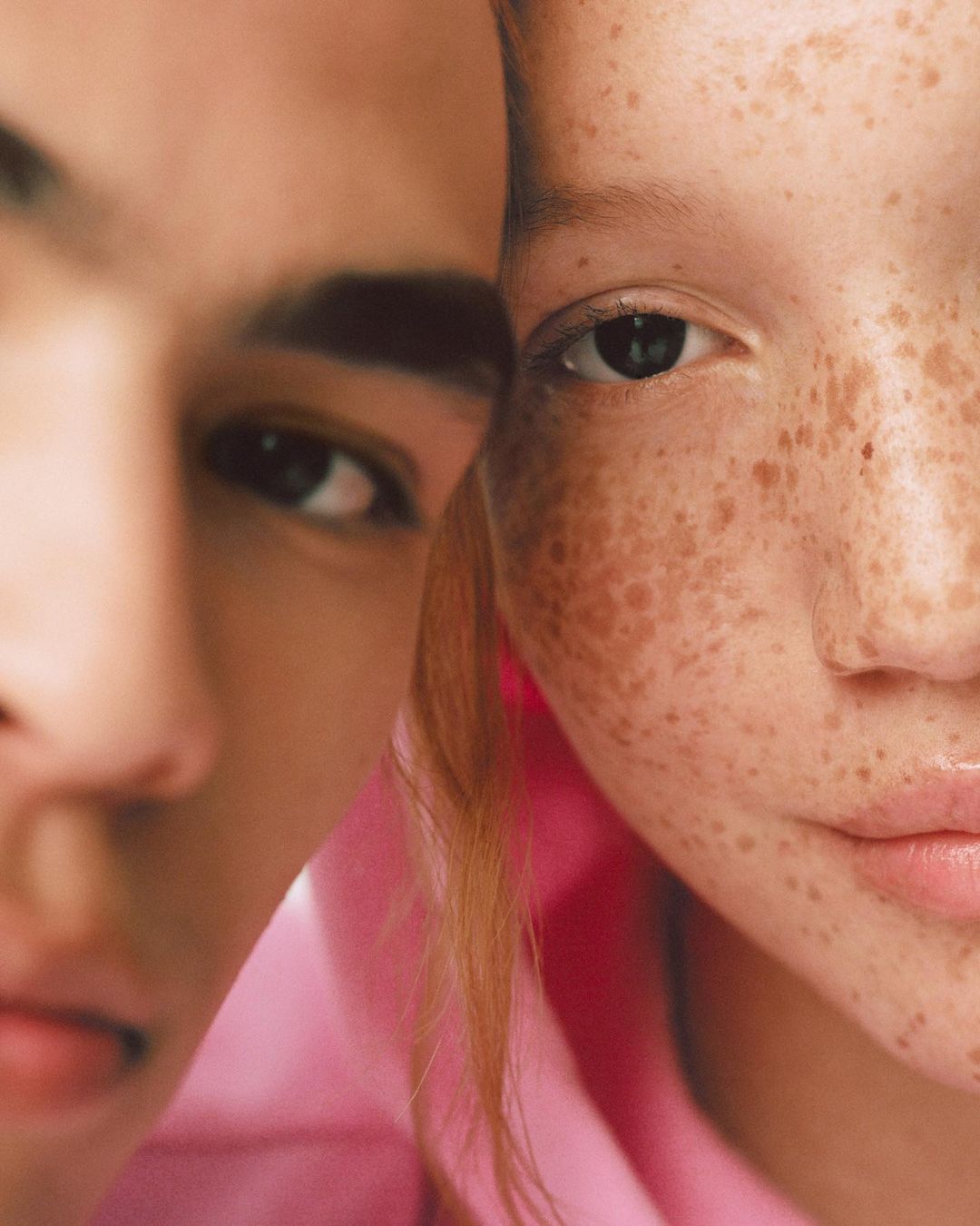 Close-up studio portrait of two young adult models, one male and one female, posing closely together with a solid pink background. 