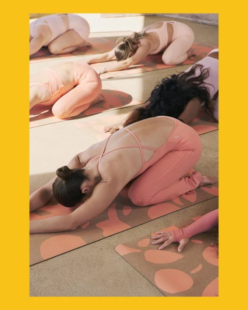 Group of women in pink athleisure wear performing child's pose on yoga mats in a bright indoor space with natural light.