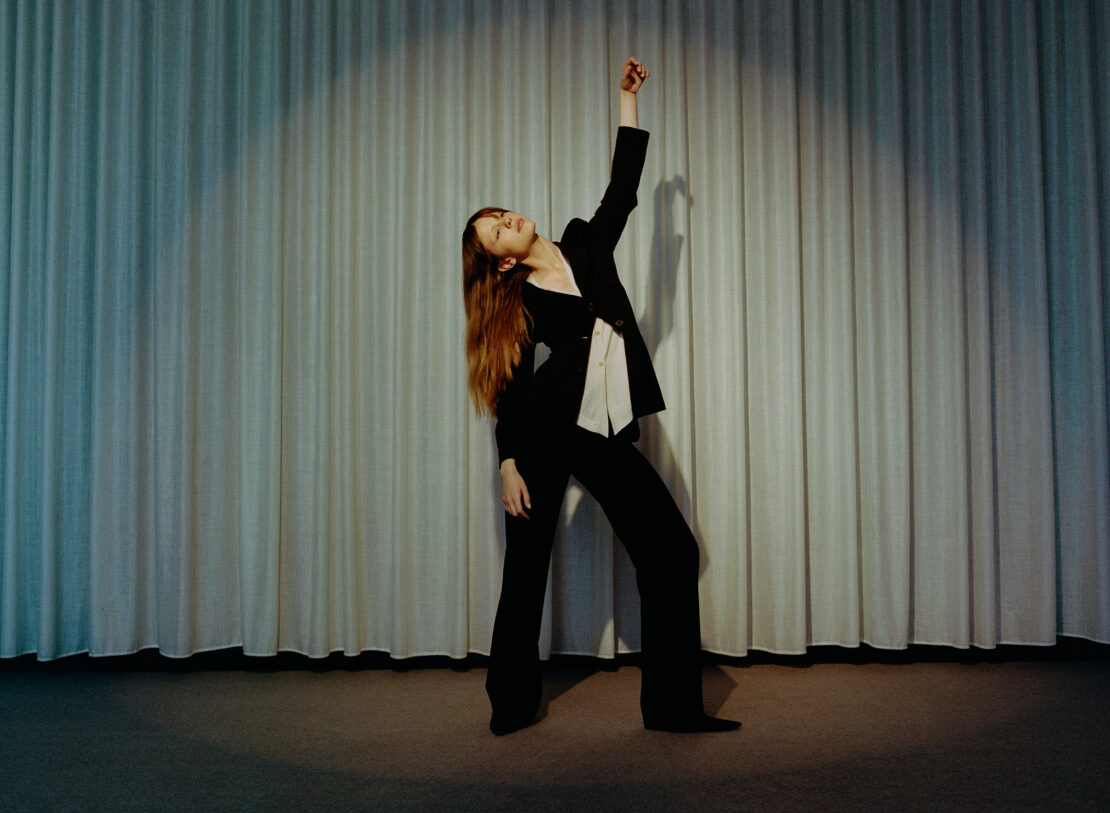 A woman in a black suit poses in front of a grey curtain, illuminated by a spotlight, in a portrait with her arm raised and head tilted back.