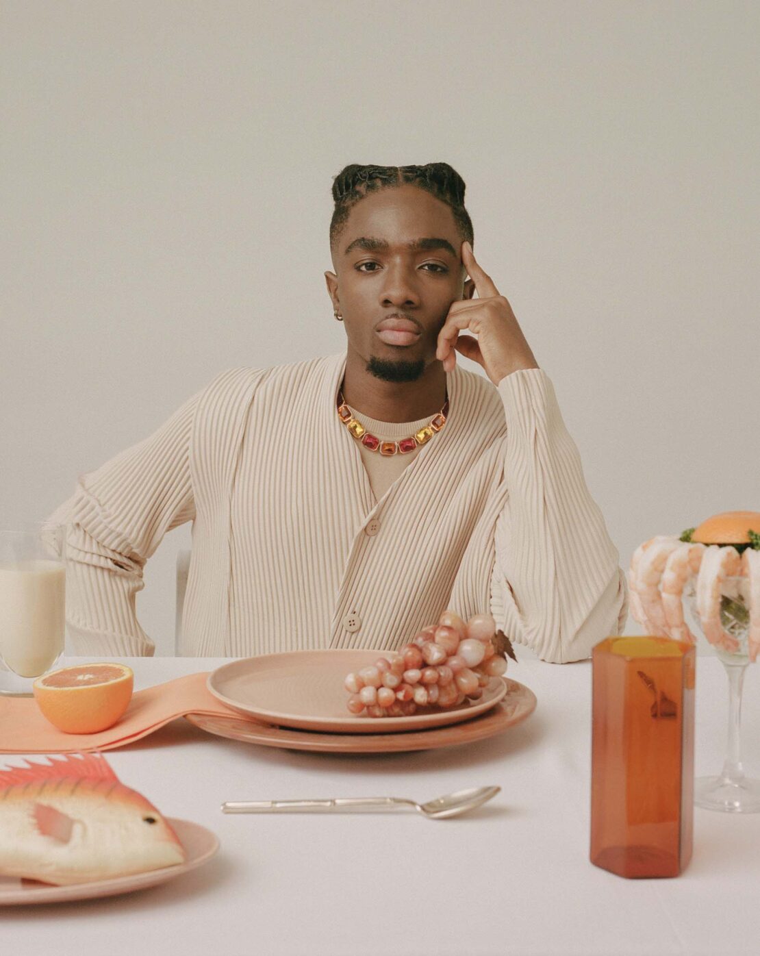 Portrait of a man at a table with grapes, a plate, and a fish ornament. He wears a beige shirt and colorful necklace.