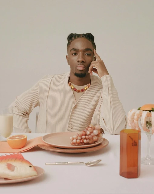 Portrait of a man at a table with grapes, a plate, and a fish ornament. He wears a beige shirt and colorful necklace.