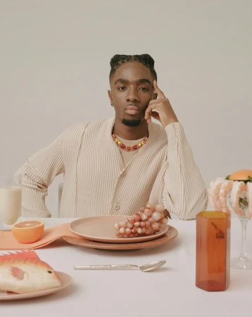 Portrait of a man at a table with grapes, a plate, and a fish ornament. He wears a beige shirt and colorful necklace.