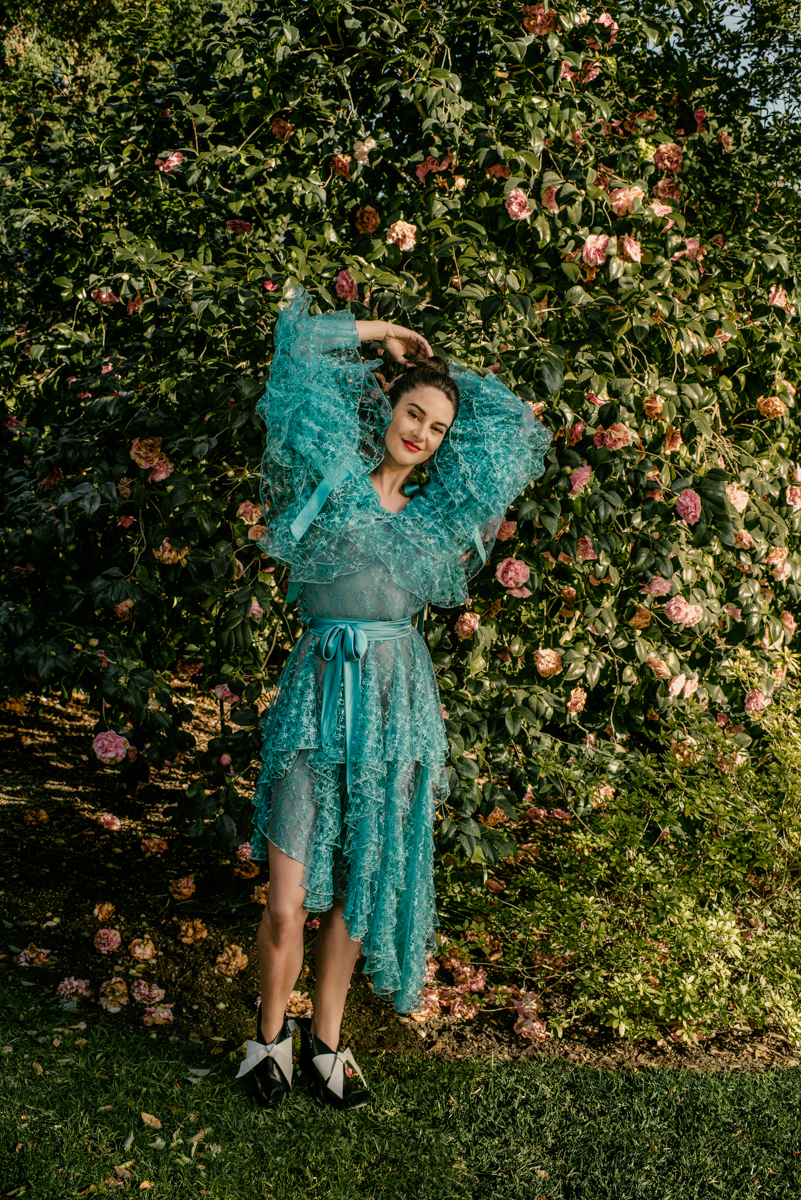 An adult woman poses in a semi-sheer turquoise dress, black-and-white shoes, and an elaborate ruffled top in a verdant outdoor garden setting.