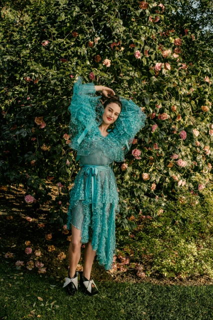 An adult woman poses in a semi-sheer turquoise dress, black-and-white shoes, and an elaborate ruffled top in a verdant outdoor garden setting.