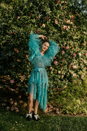 An adult woman poses in a semi-sheer turquoise dress, black-and-white shoes, and an elaborate ruffled top in a verdant outdoor garden setting.