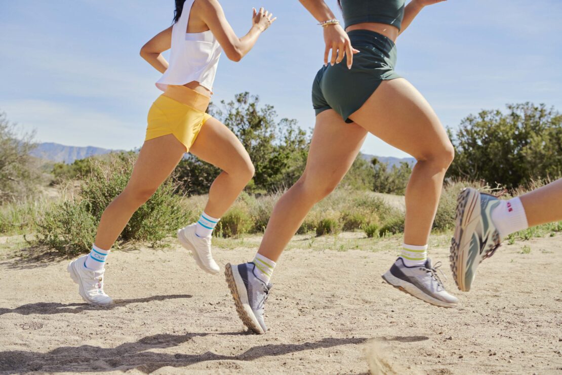 Two women are running on a dirt path with mountains in the background. They are wearing athletic wear and running shoes.