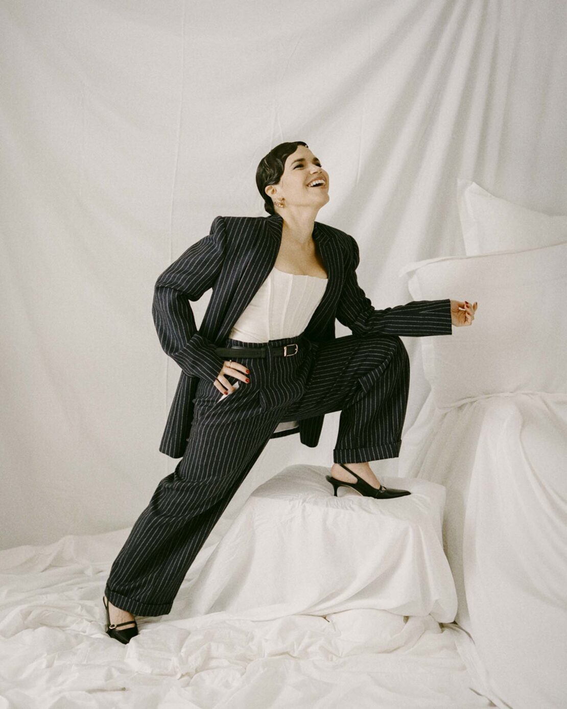 A smiling woman in a pinstripe suit with a white corset top poses dynamically on white sheets in a studio setting, exuding confidence.