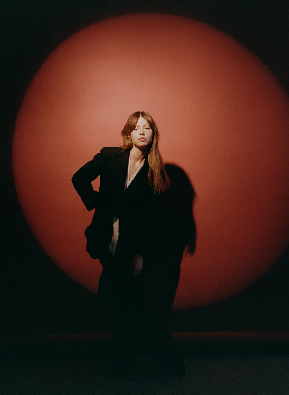 Portrait of a young woman in a black jacket with a red spotlight background, looking at the camera with a neutral expression.