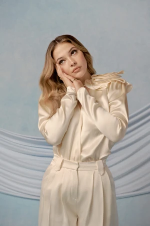Young woman with blonde wavy hair poses in an off-white satin blouse and wide-legged pants in front of a draped blue background.