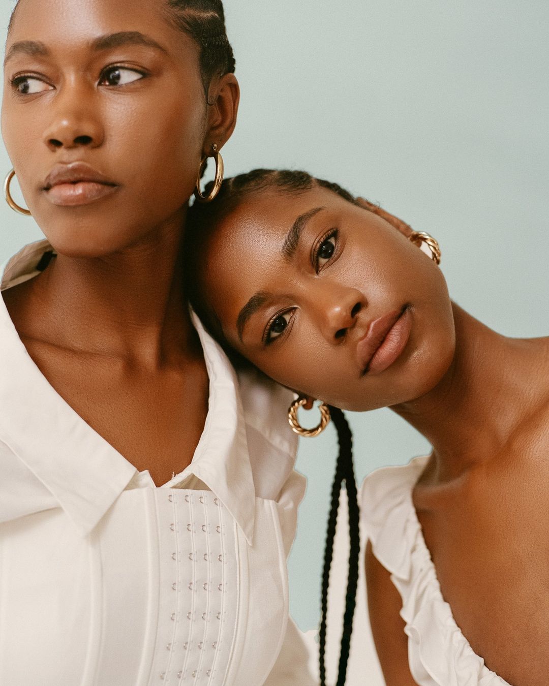 Two adult women with braided hair pose indoors wearing white tops, and gold hoop earrings against a light blue background.