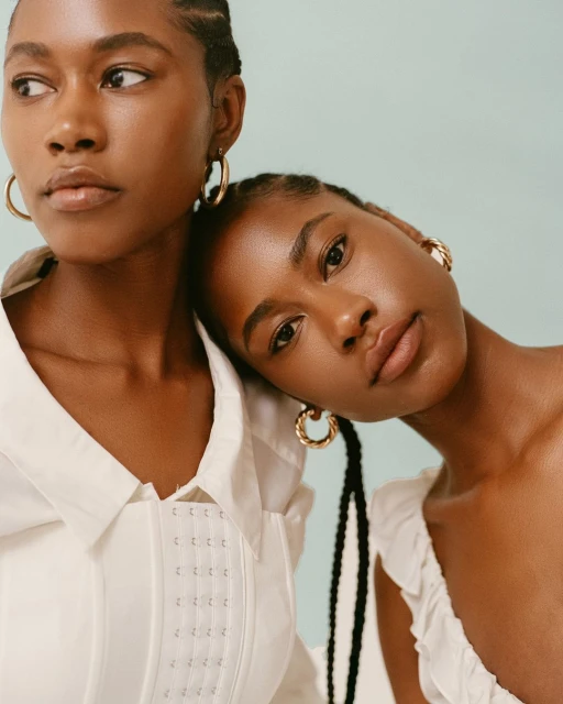 Two adult women with braided hair pose indoors wearing white tops, and gold hoop earrings against a light blue background.