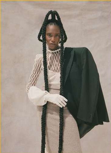 Elegant woman with long braided hairstyle and black jacket poses against a neutral backdrop, showcasing a blend of fashion and sophistication.