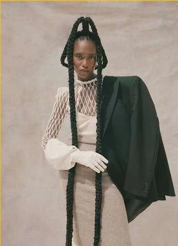 Elegant woman with long braided hairstyle and black jacket poses against a neutral backdrop, showcasing a blend of fashion and sophistication.