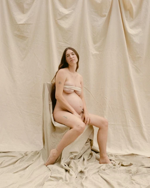 Pregnant woman sits nude on a draped chair in front of a fabric backdrop. Her long brown hair falls past her shoulders.