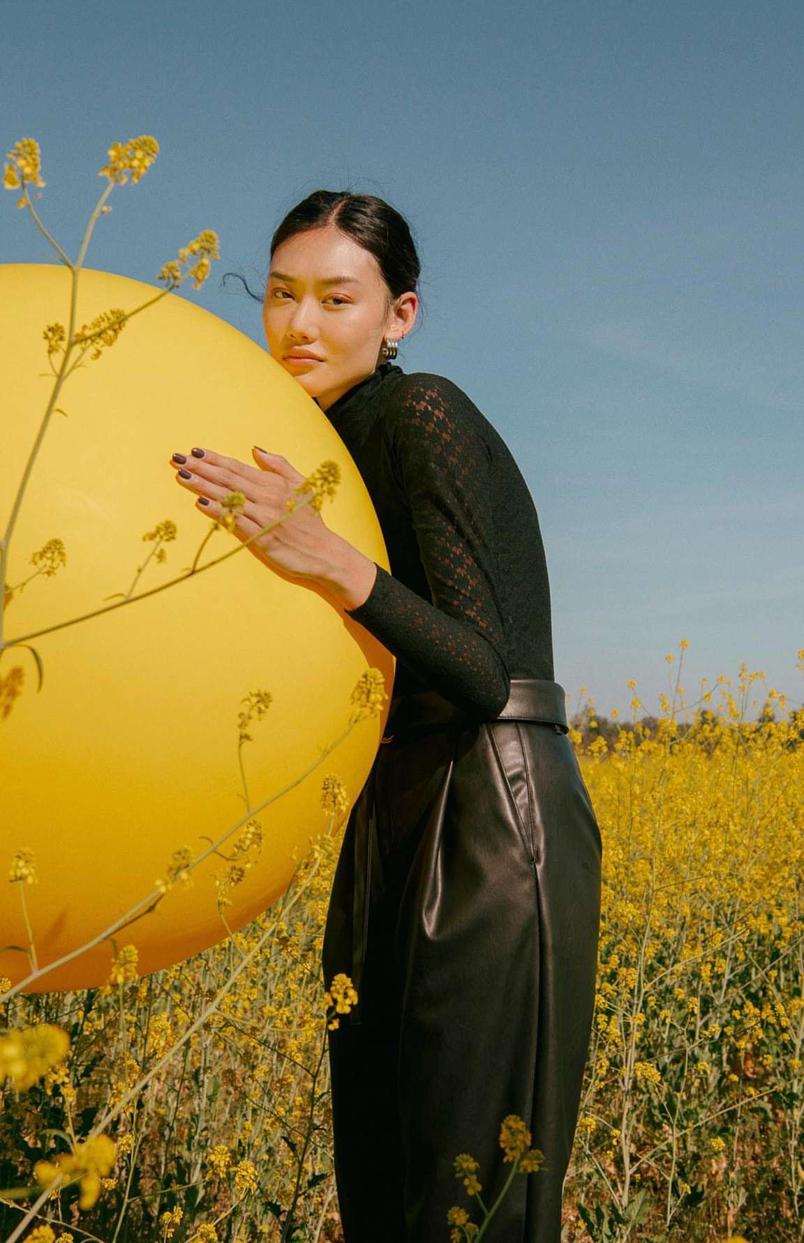 Young woman in black hugging a yellow balloon in a field of yellow wildflowers under a blue sky in a medium portrait shot.