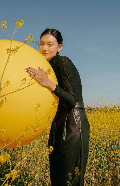 Young woman in black hugging a yellow balloon in a field of yellow wildflowers under a blue sky in a medium portrait shot.