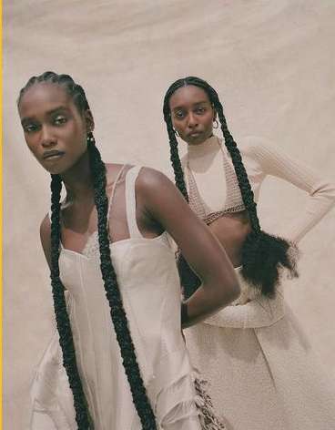 Two dark-skinned women pose in cream-colored dresses with long braids against a neutral backdrop in a studio fashion editorial shot.