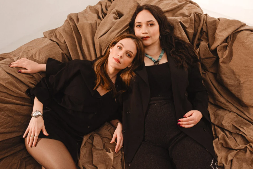Two adult women in black outfits pose indoors on a brown textured surface, captured in a medium shot with a neutral facial expression.