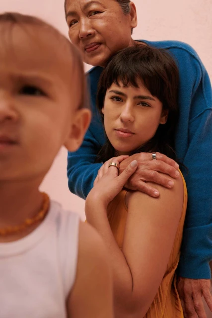 Three generations of women pose indoors with a pink background, an infant, a young adult, and an older woman; shot in portrait orientation.