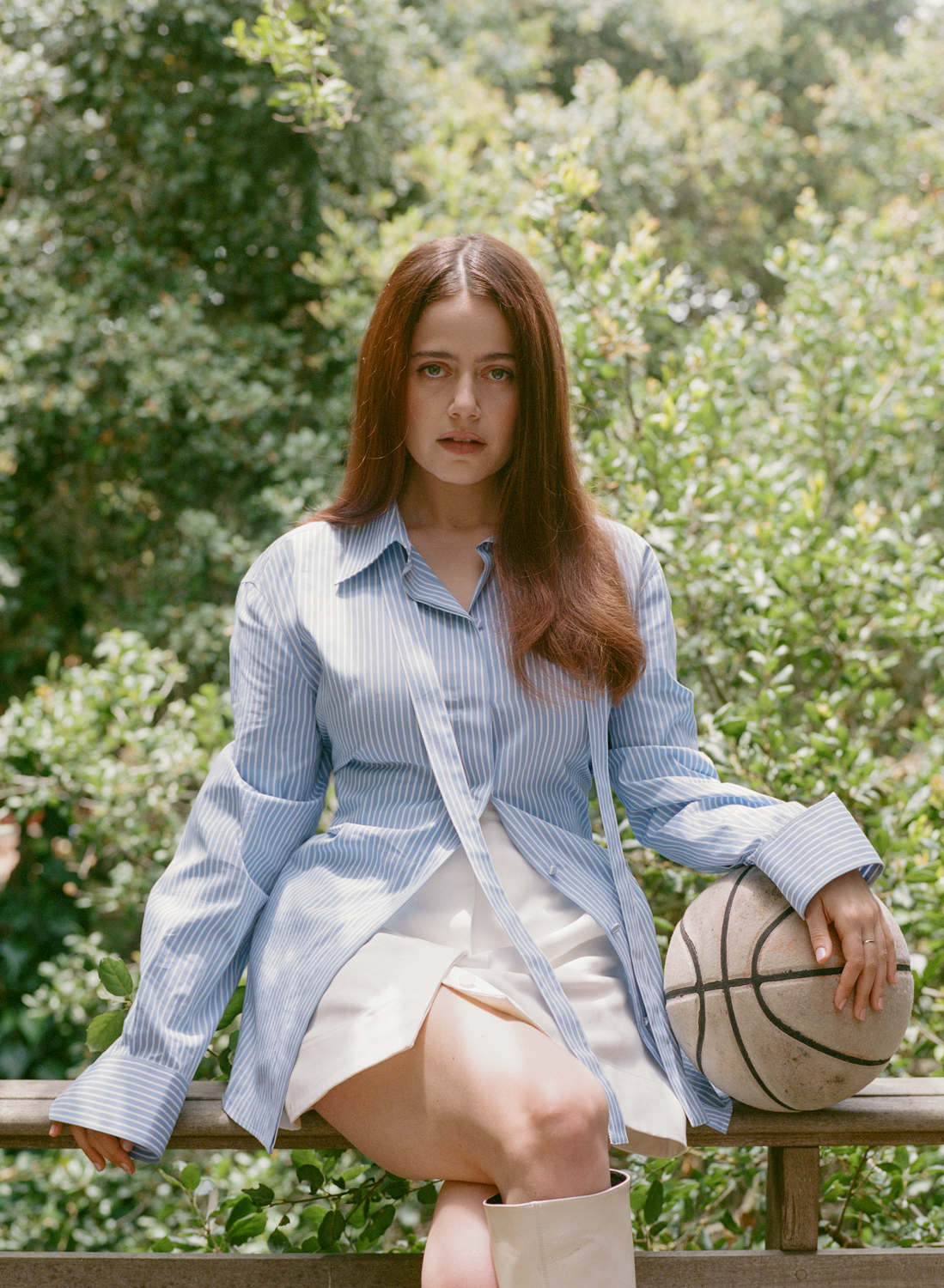 A fair-skinned adult female with long auburn hair sits on a brown wooden rail, holding a basketball, with a green leafy background.