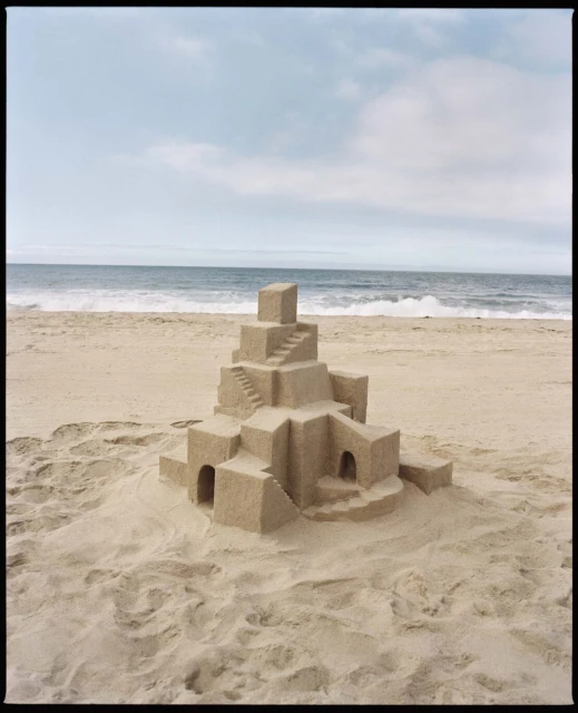 A sandcastle on a sandy beach under a blue sky with clouds and ocean waves in the background on a sunny day.