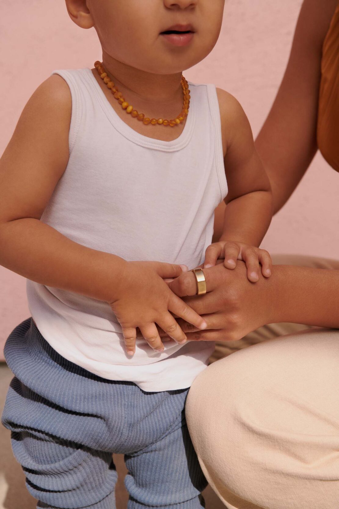 Close-up portrait of a toddler wearing a white tank top and blue textured pants standing next to an adult holding hands.