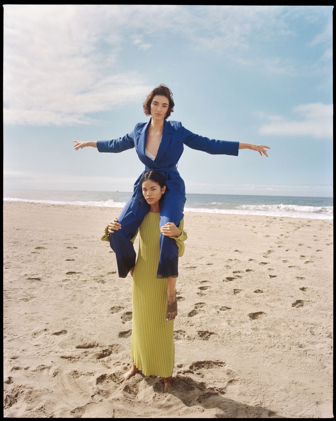Two women pose on a sandy beach, one giving the other a piggyback ride. The woman on top wears a blue suit with the ocean in the background.